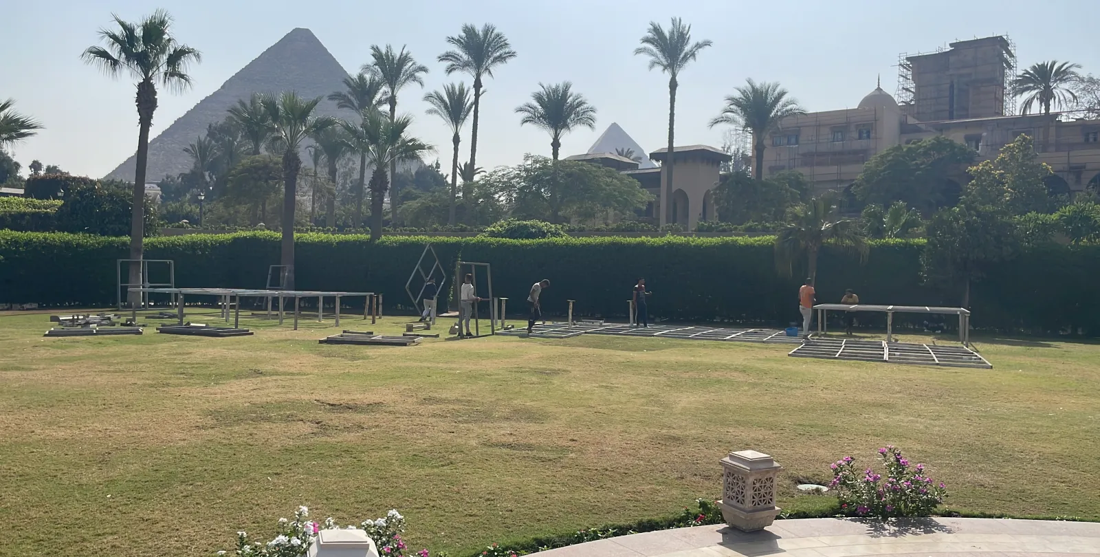 An empty hotel lawn with a few truss sections laid down, palms and the Great Pyramid behind.
