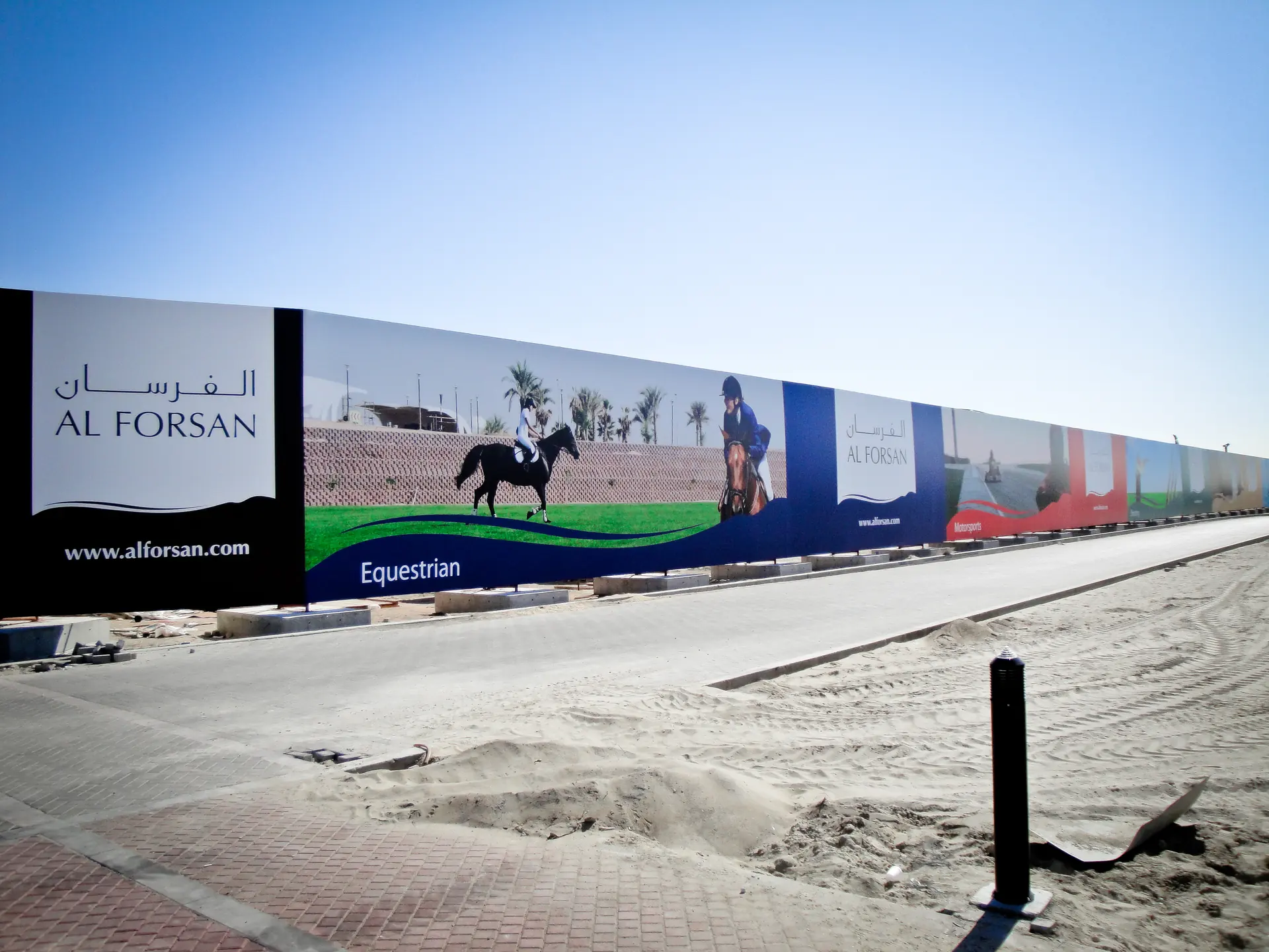 Photograph: the hoarding along the road, a black panel with the two names, then riders on a blue wave marked Equestrian
