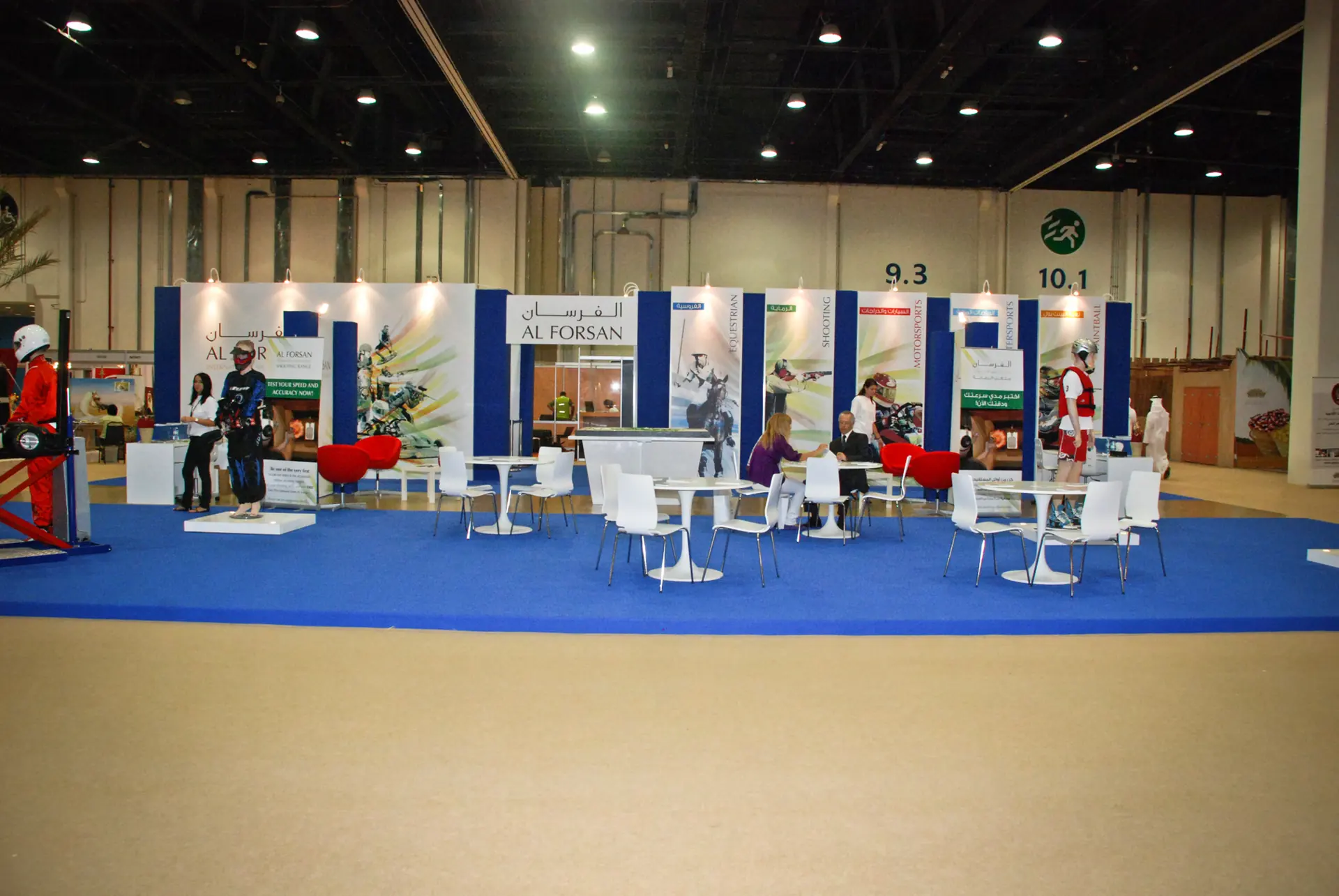 Photograph: the Al Forsan exhibition stand on a blue carpet, white walls with the sports, tables and chairs in front
