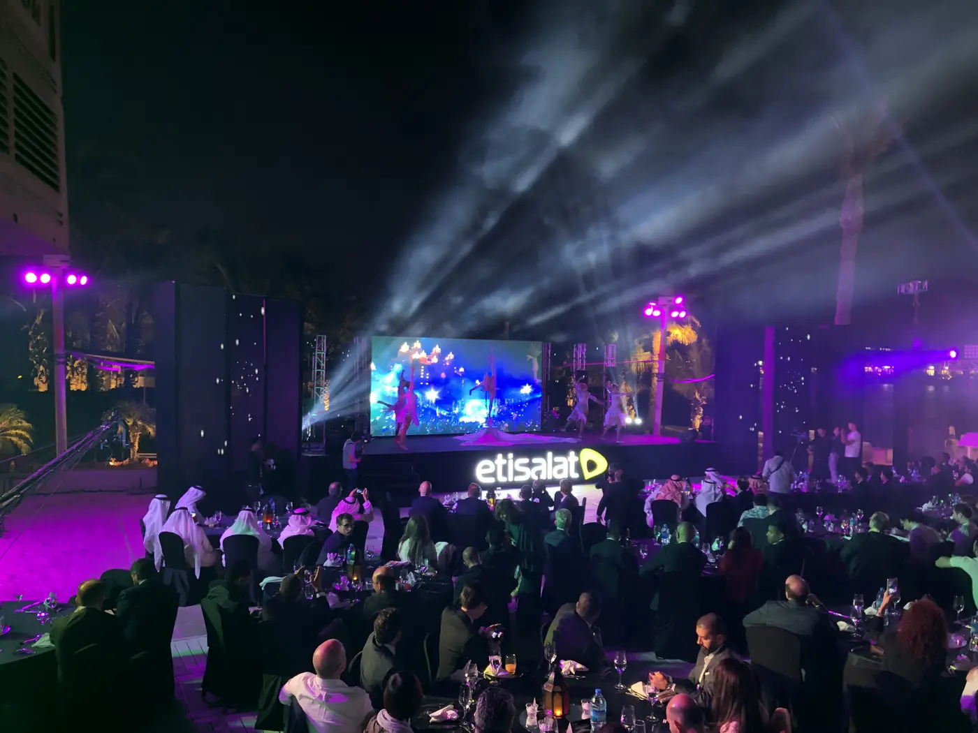 Photograph at night: the full room of tables, the stage under beams of light, the lit letters etisalat in front of it